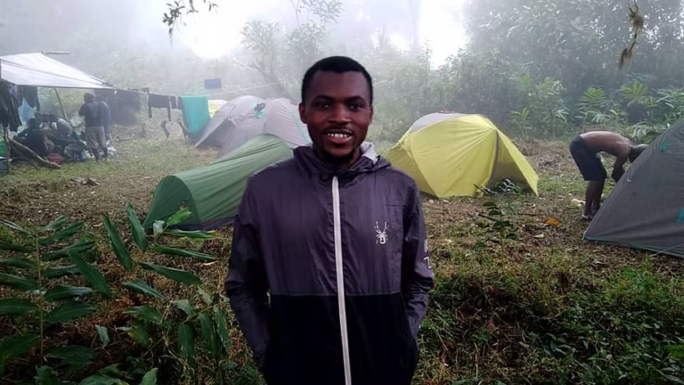 Anya Dabite Abeh standing at a forest campsite with tents behind him.