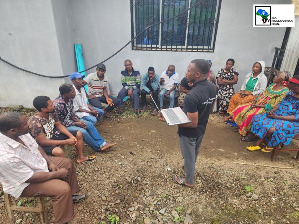 A facilitator holding a laptop while talking with seated community members outdoors.