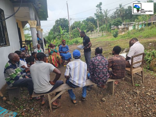 An outdoor community meeting with a facilitator addressing the seated group.