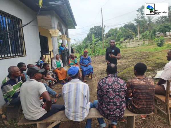A facilitator talking with community members seated in a semicircle outdoors.
