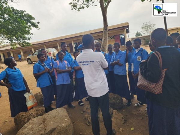Students in blue uniforms listening to a facilitator in a schoolyard.