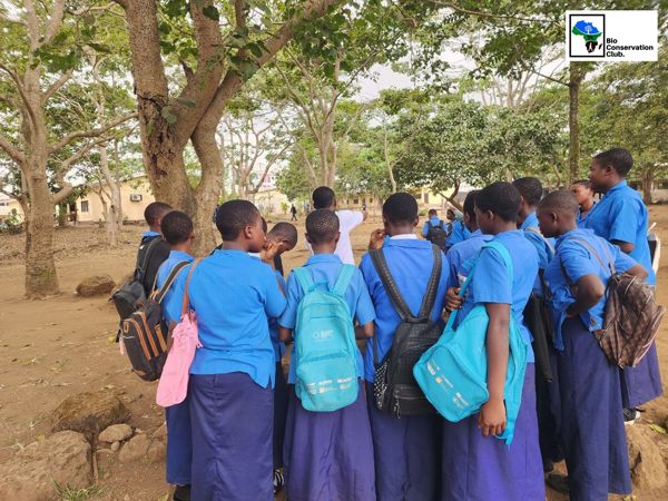 Students gathered around a facilitator under trees in a schoolyard.