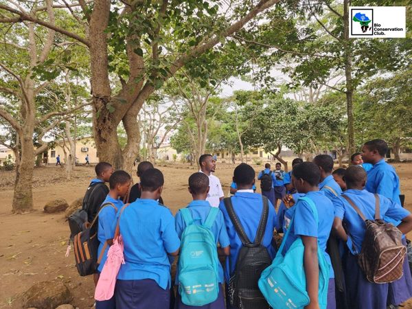 A facilitator speaking with a group of students beneath schoolyard trees.